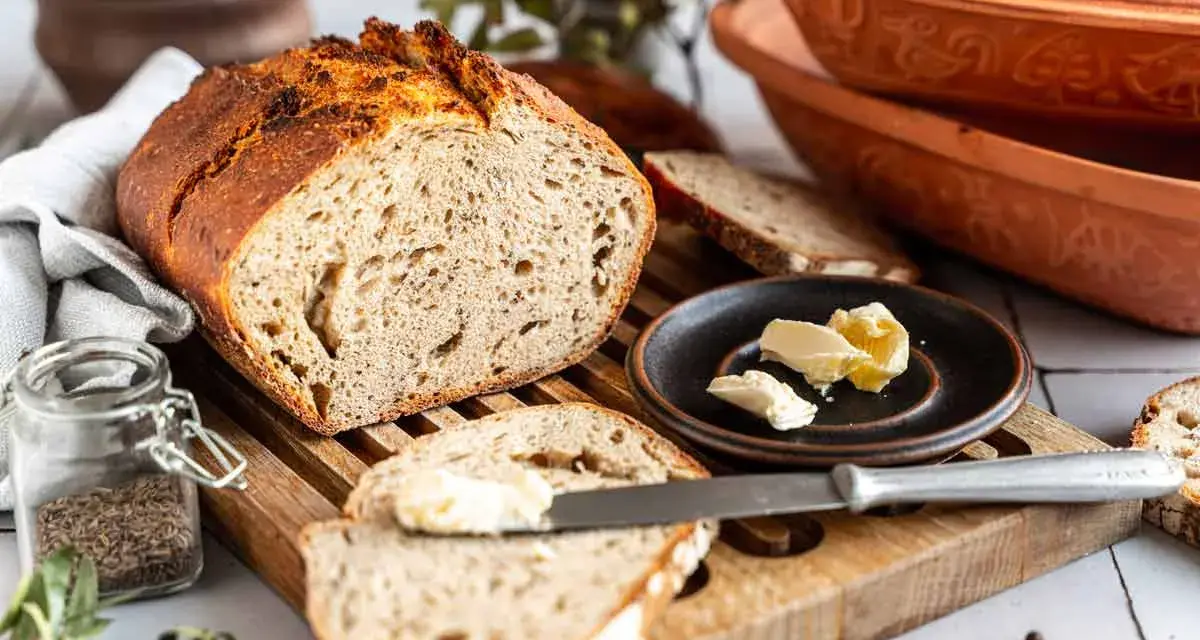 Ein rustikales Roggenbrot mit Sauerteig, aufgeschnitten und mit Butter bestrichen. Daneben ein Glas Kümmel und ein Messer.