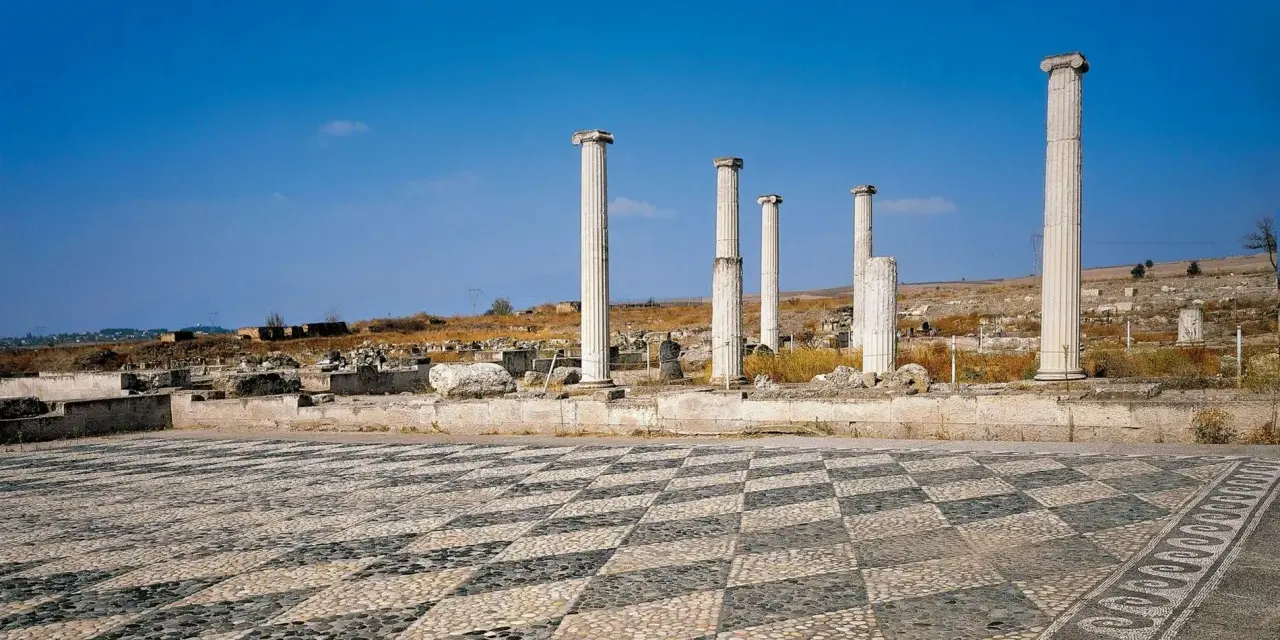 Ruines antiques avec colonnes imposantes et un sol en mosaïque de galets, évoquant l'origine d'Alexandre le Grand.