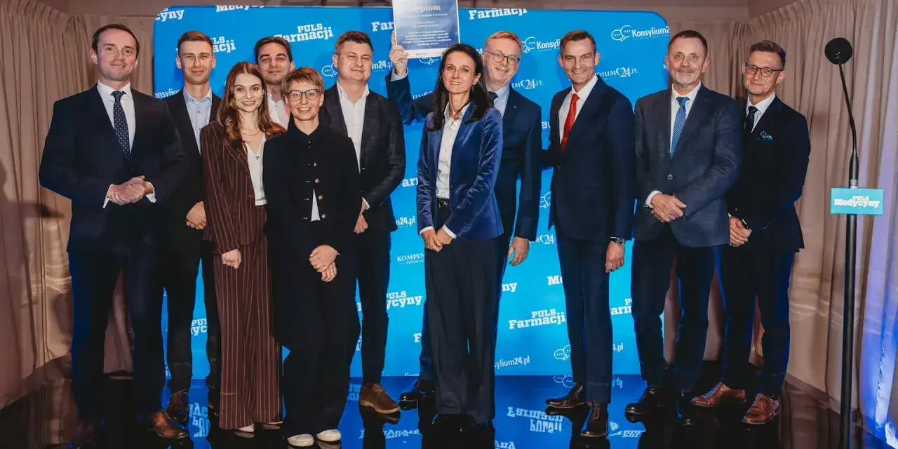 Grupa ludzi na scenie, wśród nich Janusz Sowa, odbiera dyplom. W tle niebieskie banery z logo Puls Farmacji.