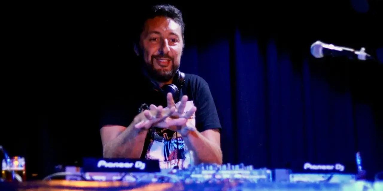 Un dj hip hop souriant, mains en l'air, mixe sur scène devant un public invisible.