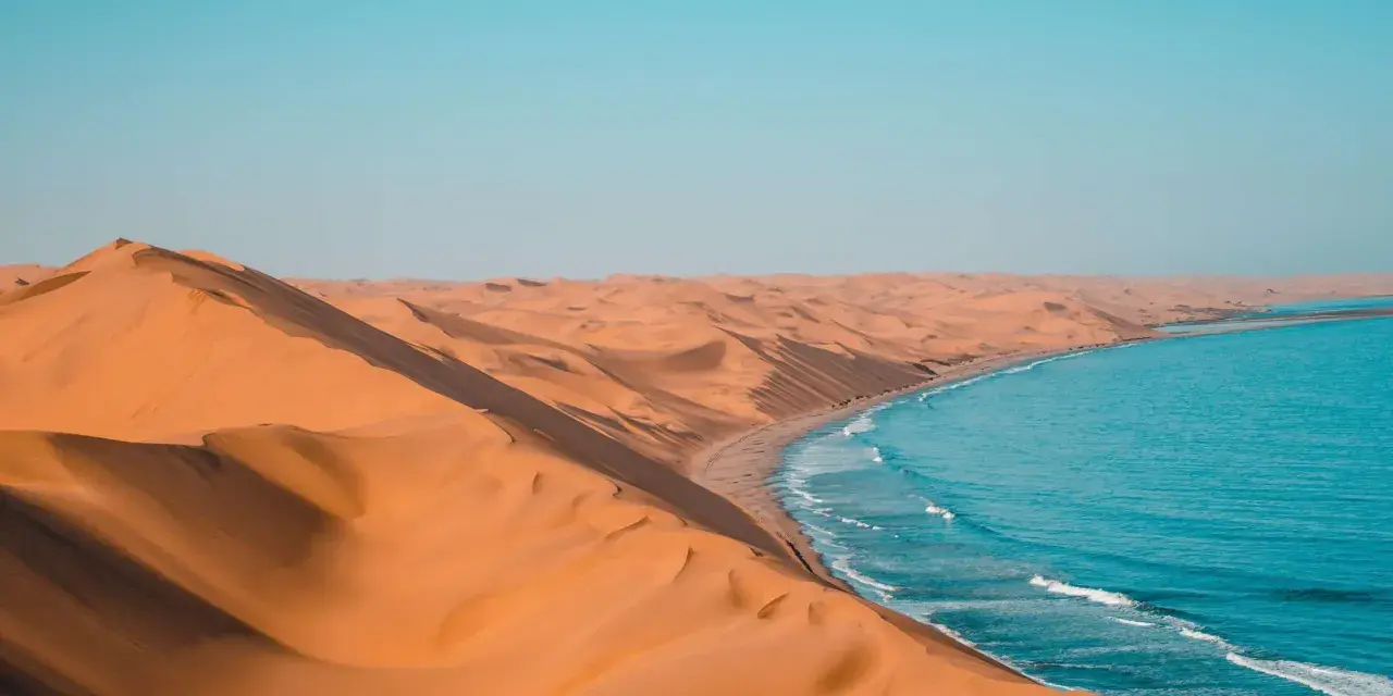 Piaskowe wydmy Namibii spotykają się z turkusowym oceanem. Idealna namibia pogoda na podziwianie tego kontrastu.