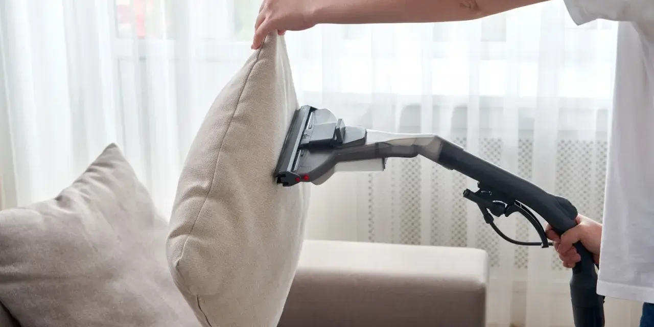 En person använder en soffdammsugare för att rengöra en ljusbeige kudde på en soffa.