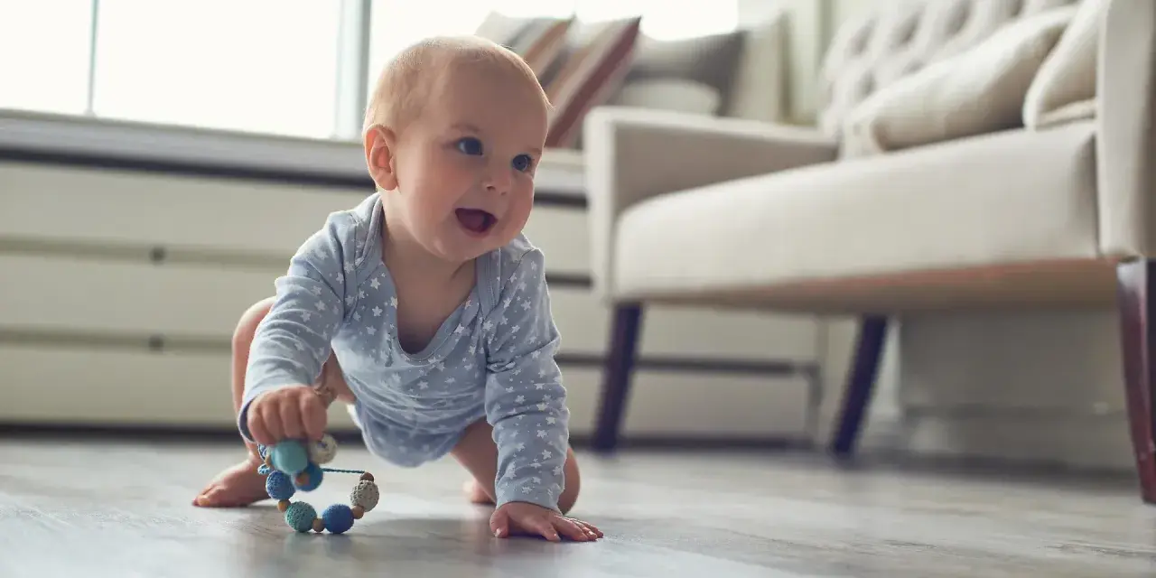 Ein Baby in blauem Sternen-Body krabbelt fröhlich über den Holzboden und greift nach einer bunten Rassel. So erkunden Babys die Welt!
