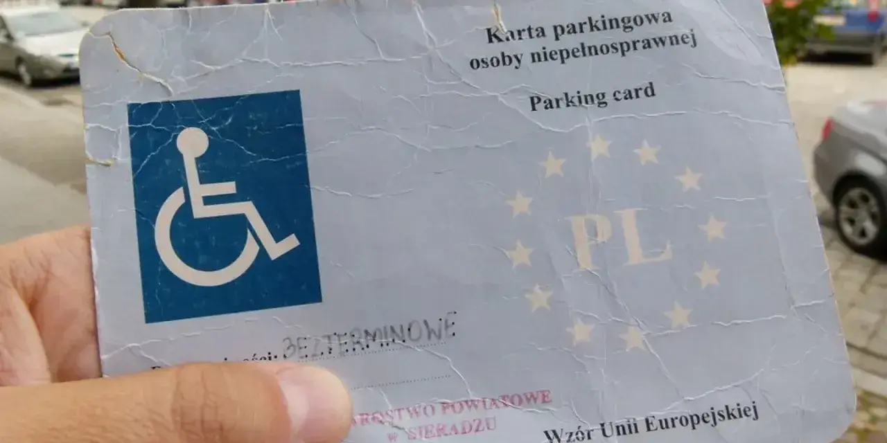 Karta parkingowa dla osoby z niepełnosprawnością, stopień umiarkowany. W tle samochody i budynki.