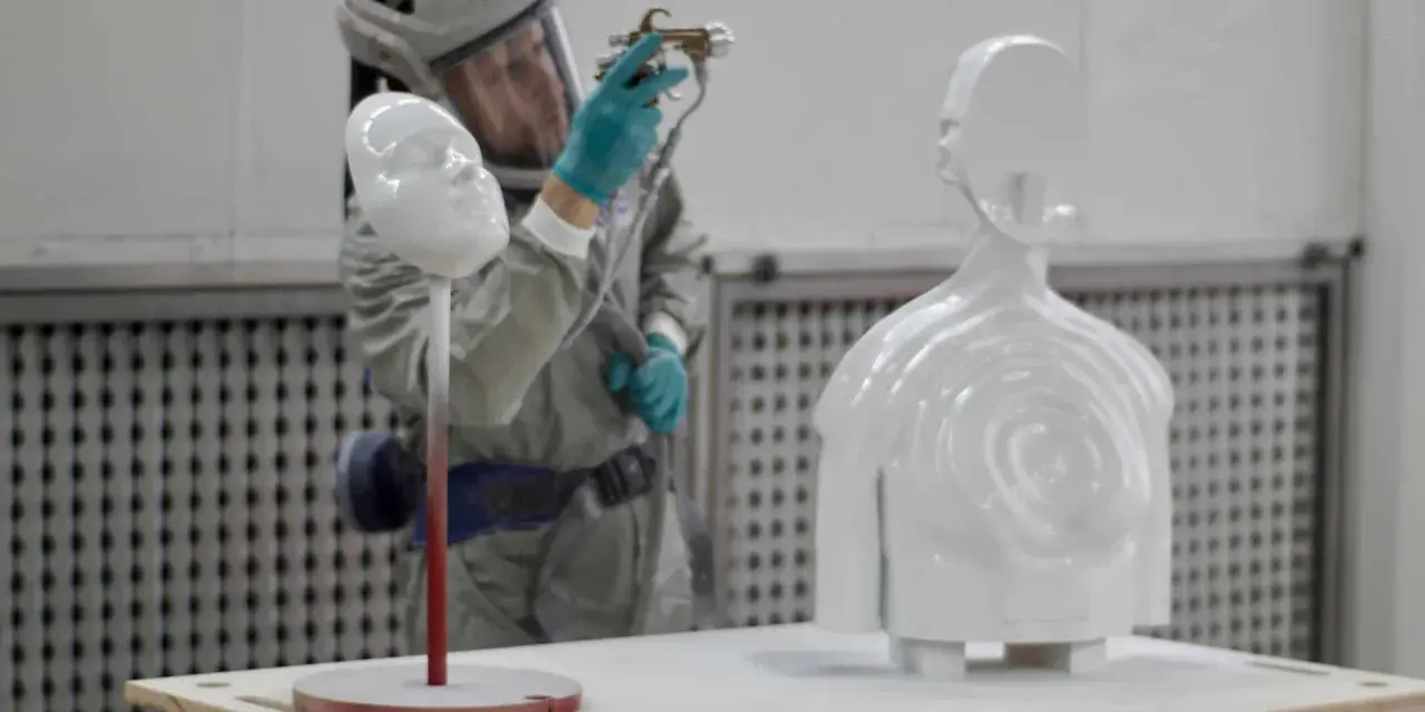 A person in a protective suit sprays a white 3D printed bust and torso, demonstrating how to paint 3d prints for a smooth finish.