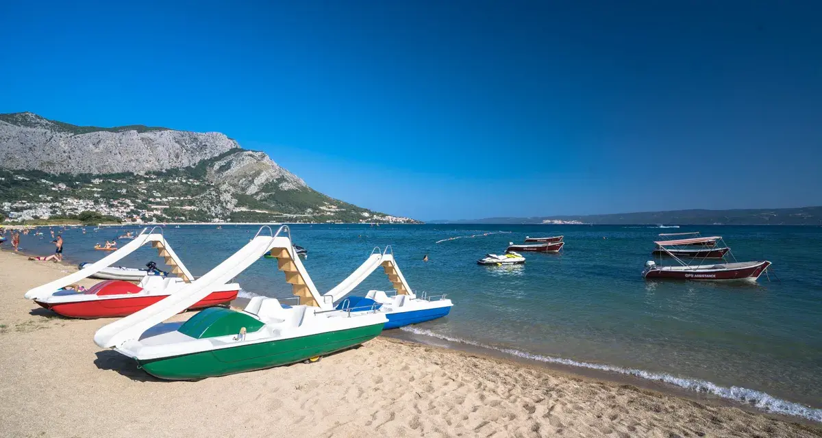 Plaża z łódkami i zjeżdżalniami, idealna na wakacje w camping Galeb. W tle góry i turkusowe morze.