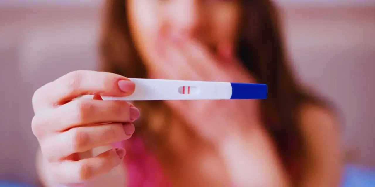 Dwa czerwone paski na teście ciążowym. Kobieta z zakrytymi ustami, być może planuje zwolnienie lekarskie w ciąży.