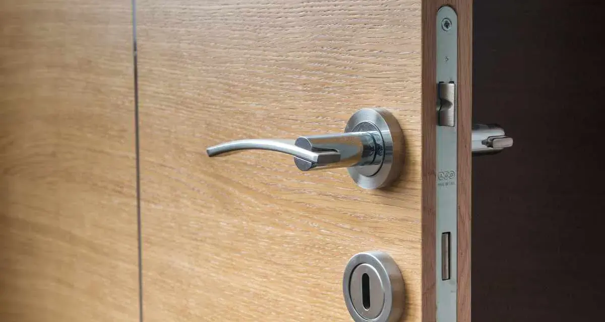 Maniglia e serratura di una porta in legno chiaro, con un'ombra scura che suggerisce una porta che cigola.