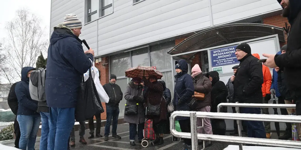 Grupa ludzi stoi przed budynkiem spółdzielni mieszkaniowej w deszczu, niektórzy pod parasolem.