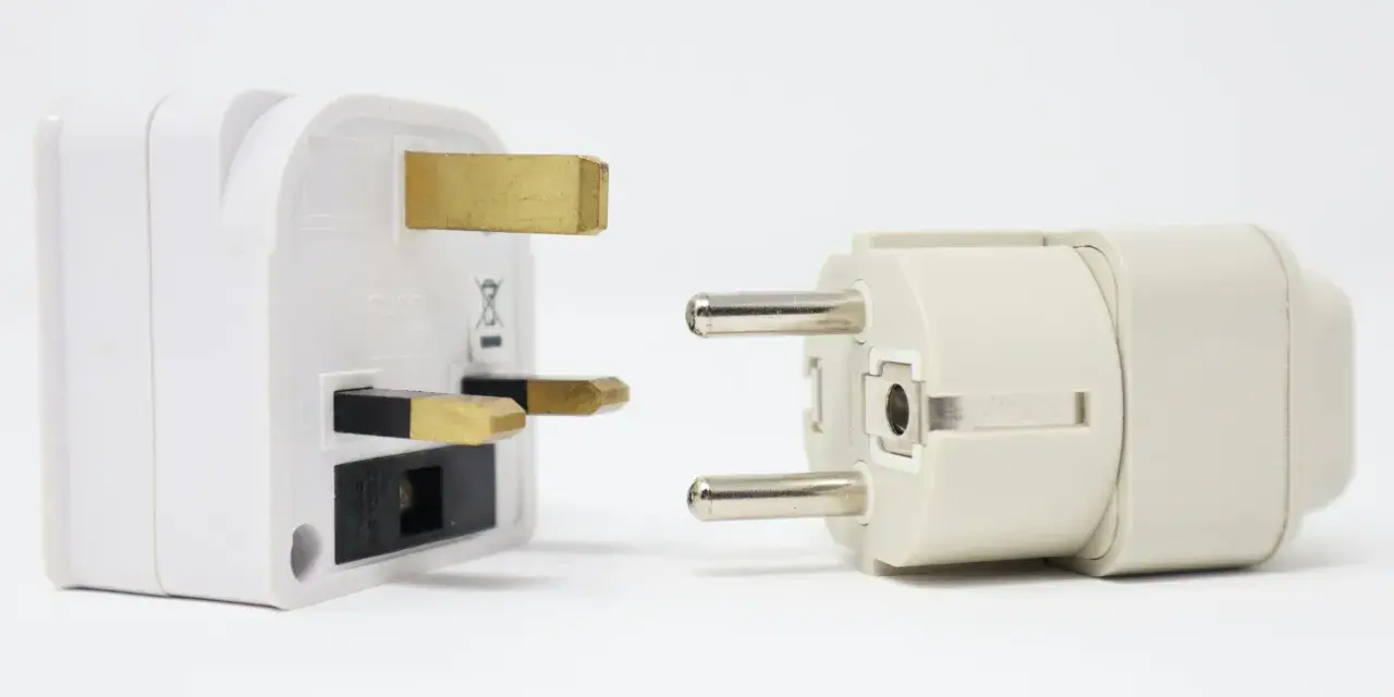 Adaptadores para diferentes tipos de enchufes: uno blanco con tres clavijas doradas y otro beige con dos clavijas plateadas.