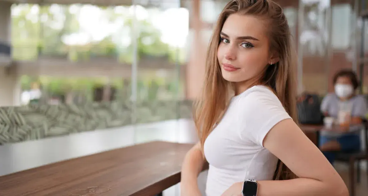 Uśmiechnięta blondynka w białej bluzce, z zegarkiem na ręku, siedzi przy stoliku. Czy to jej sekret, jak uwieść faceta?