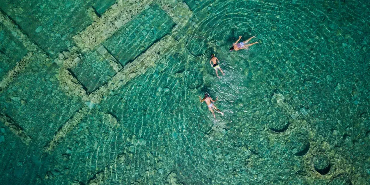 Swimmers explore the sunken city of Epidaurus, its ancient stone structures visible beneath the clear turquoise water.