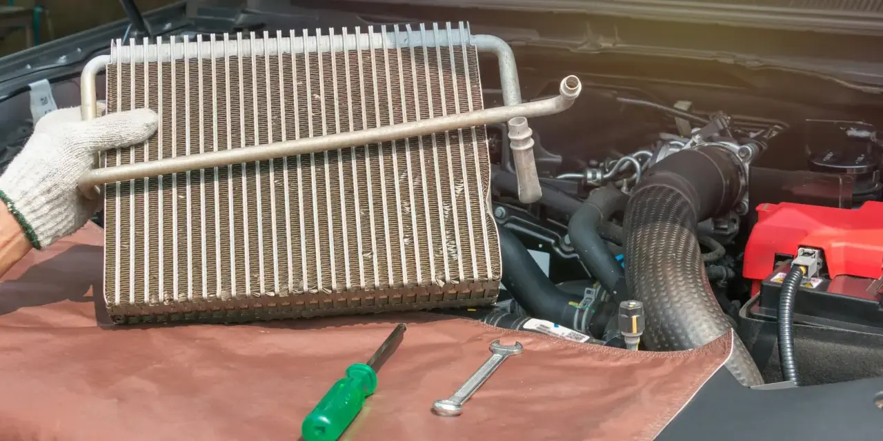 Gloved hand holds car AC evaporator. Tools and engine parts visible. Learn how to make AC colder in car by fixing this component.