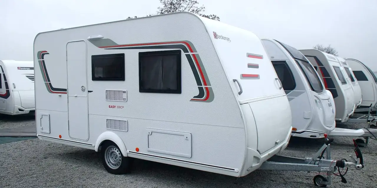 Biała przyczepa kempingowa Sterckeman Easy 390CP z czerwono-szarymi akcentami, zaparkowana na żwirowisku obok innych przyczep.