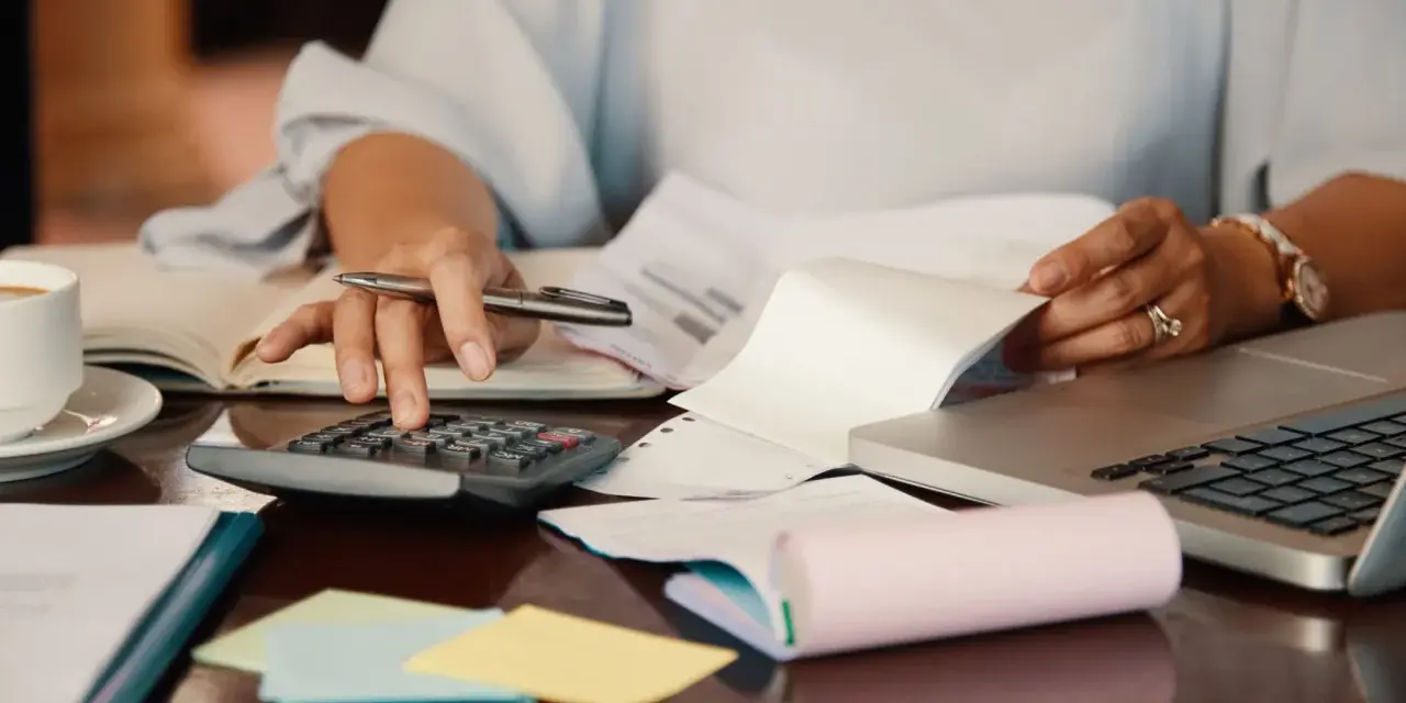 Persona revisa documentos y usa calculadora para el modelo 184. Hay café, portátil y notas adhesivas.