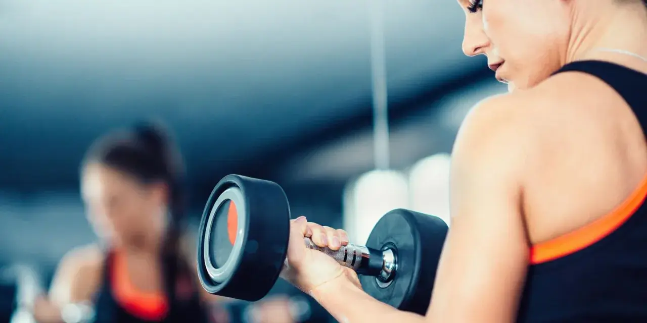 Frau hebt Hantel. Die Frage, wieviel kg darf eine Frau heben arbeitsrecht, ist hier nicht relevant, da es um Fitness geht.