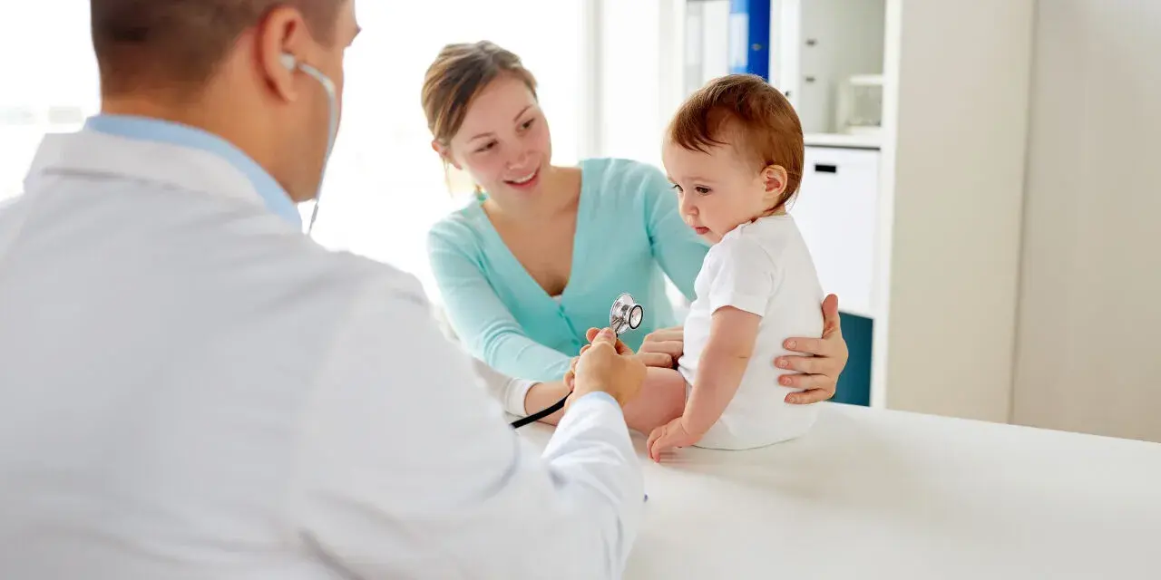 Pediatra bada niemowlę podczas pierwszej wizyty. Mama z uśmiechem obserwuje badanie.