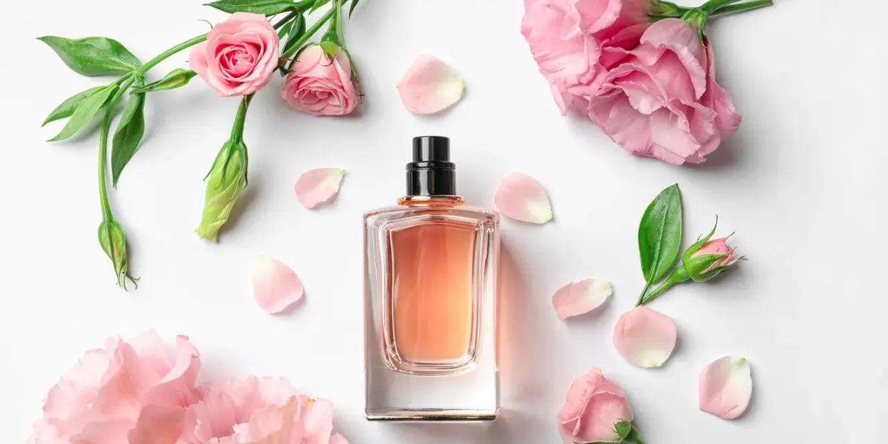 Elegancka butelka perfum z delikatnym, różowym płynem, otoczona pąkami i płatkami róż. Zapach jak słodka jeżyna.