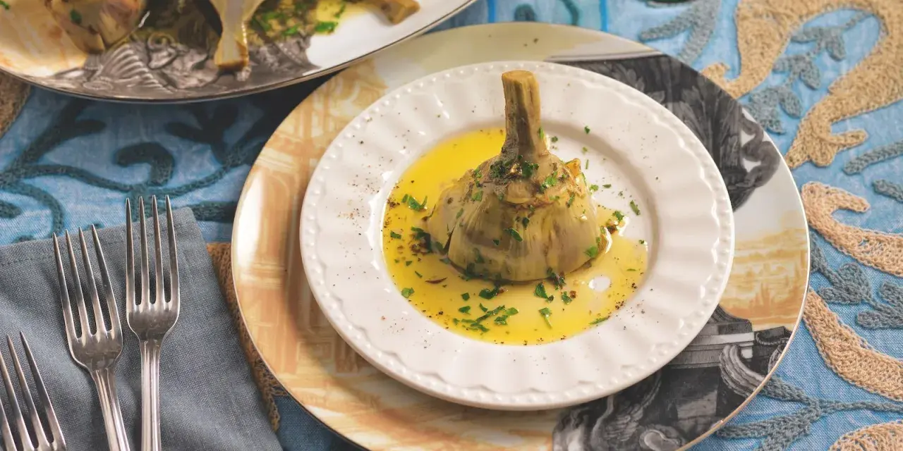 Un carciofo alla romana, tenero e profumato, adagiato su un piatto bianco con olio dorato e prezzemolo tritato.