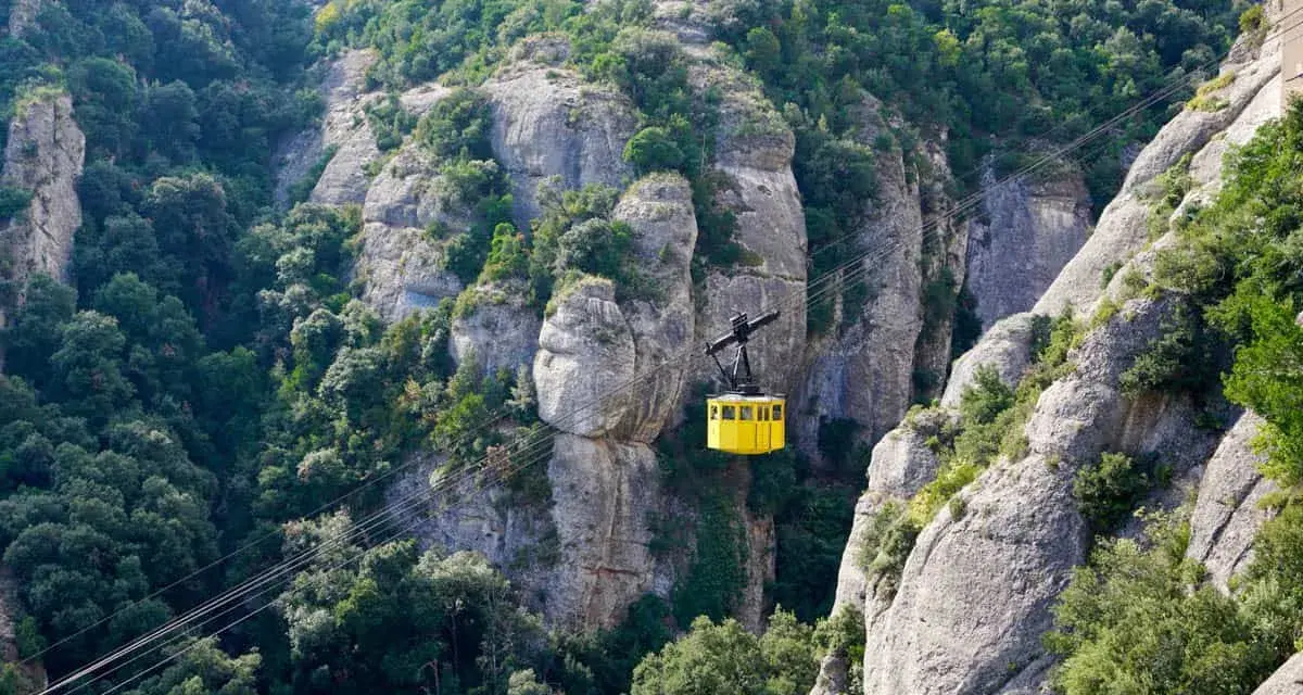 A bright yellow cable car ascends the rugged, tree-covered cliffs of Montserrat, Spain.