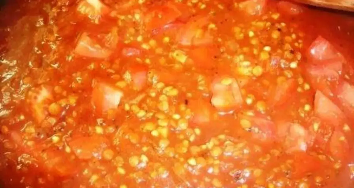 Buchweizen kochen mit Tomaten. Ein Holzlöffel rührt in der roten Sauce mit den Buchweizenkörnern und Tomatenstücken.