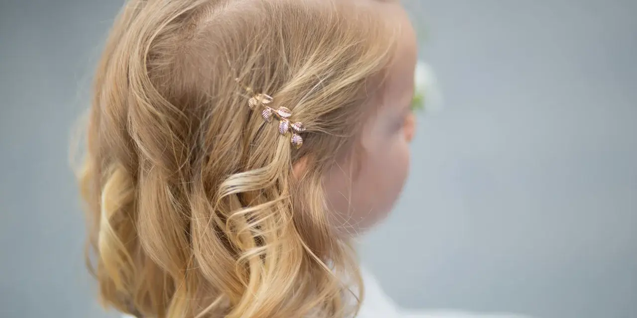 Urocze fryzury ze spinką z boku, ozdobioną złotymi listkami, dodają blasku blond włosom dziewczynki.