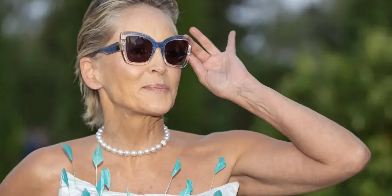 Sharon Stone, con su elegante pelo corto con canas naturales, ajusta sus gafas de sol. Lleva un collar de perlas y un vestido adornado con plumas turquesas.