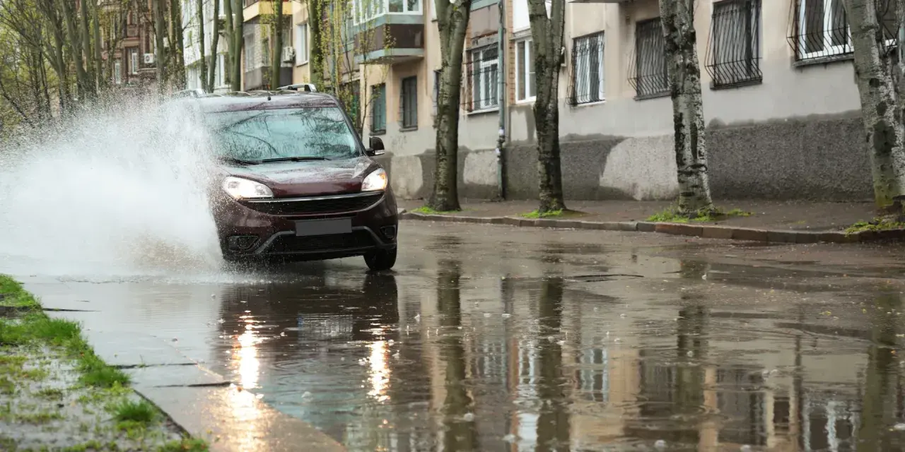 Ciemny samochód wpada w kałużę, powodując potężny **aquaplaning**. Woda rozpryskuje się na boki, a odbicia budynków i drzew tańczą na mokrej nawierzchni.