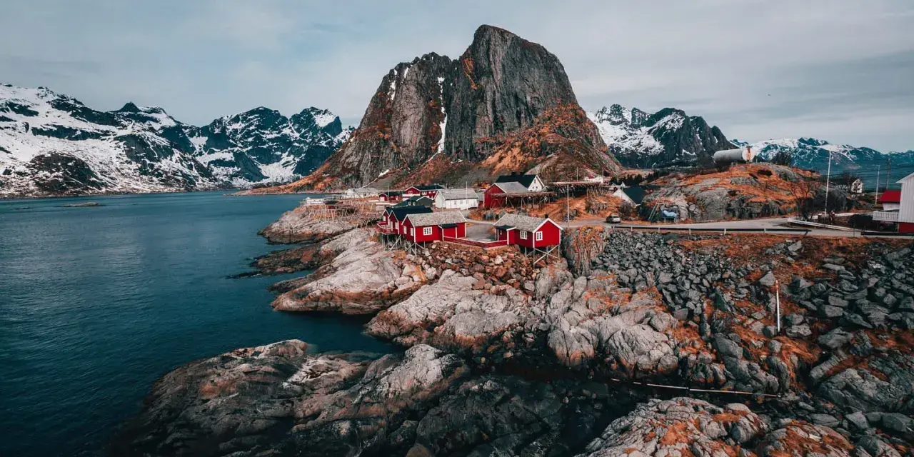 Röda fiskestugor vid en klippig kust med snöklädda berg i bakgrunden. En av de fina platserna i Norge.