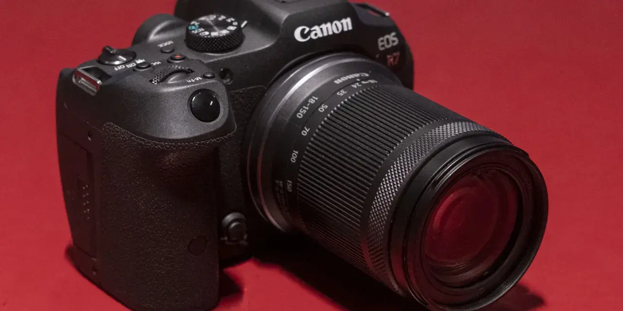 Appareil photo noir Canon EOS R7 avec objectif 18-150 mm, posé sur un fond rouge vif.