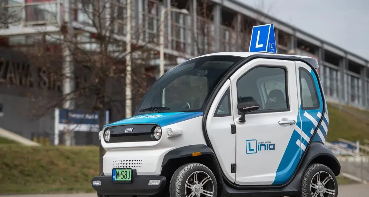Biały czterokołowiec lekki z niebieskimi akcentami, oznakowany jako pojazd nauki jazdy "Linia". Na dachu widoczny znak "L".