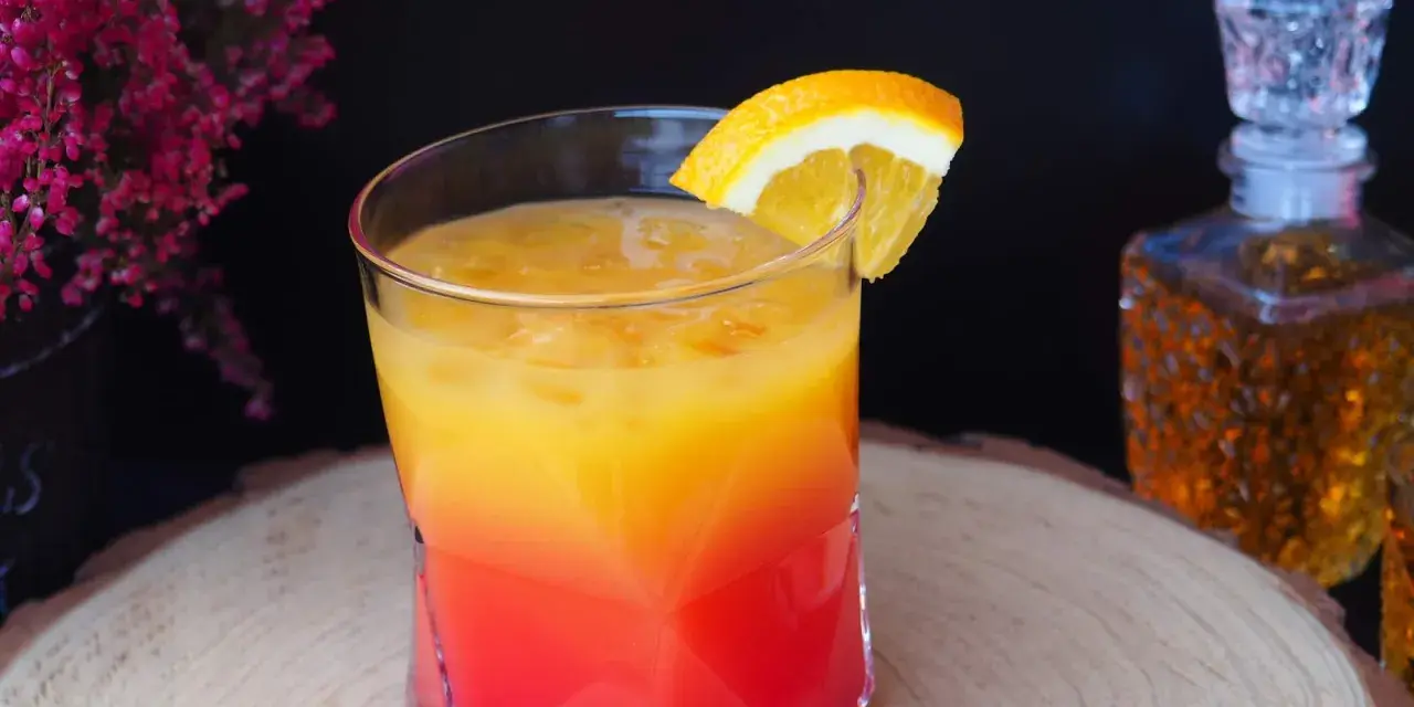 Kolorowy drink z koniakiem, ozdobiony plasterkiem pomarańczy, na tle karafki z bursztynowym alkoholem i kwitnących wrzosów.