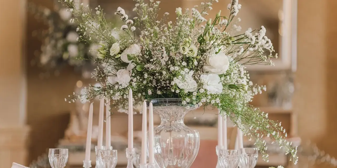Eleganckie ustawienie stołów na 40 osób. Centralny punkt stanowi bukiet białych kwiatów w szklanym wazonie, otoczony świecami i kryształowymi kieliszkami.