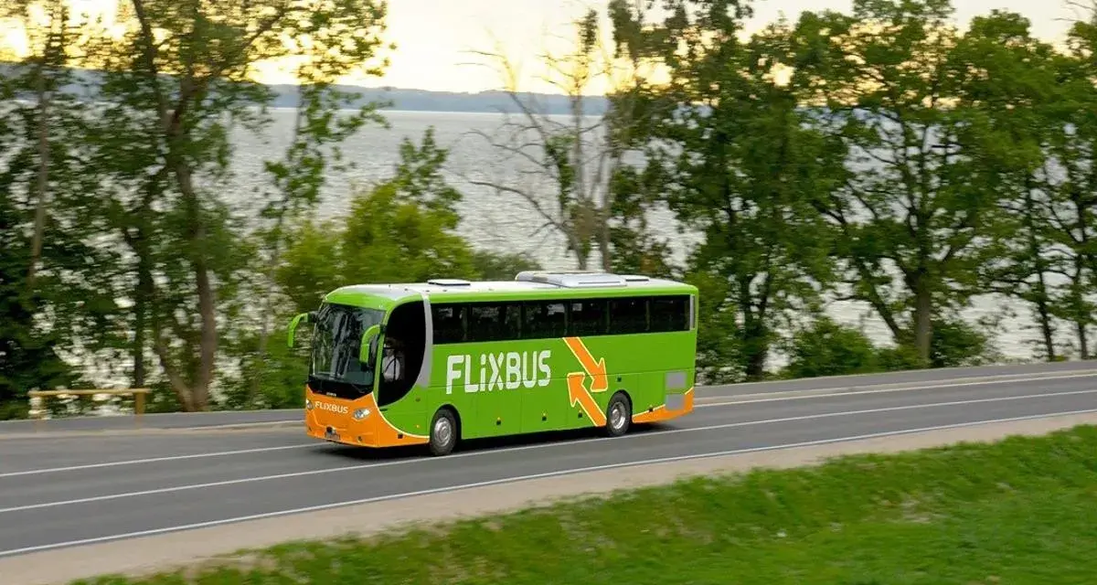 Zielony autobus Flixbus jedzie drogą przy jeziorze otoczonym drzewami.