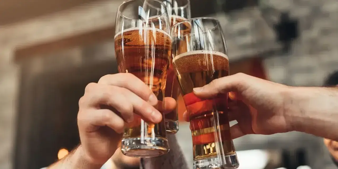 Trzy ręce unoszą kieliszki ze złotym piwem, tworząc toast. Każde piwo w szklance wygląda apetycznie.
