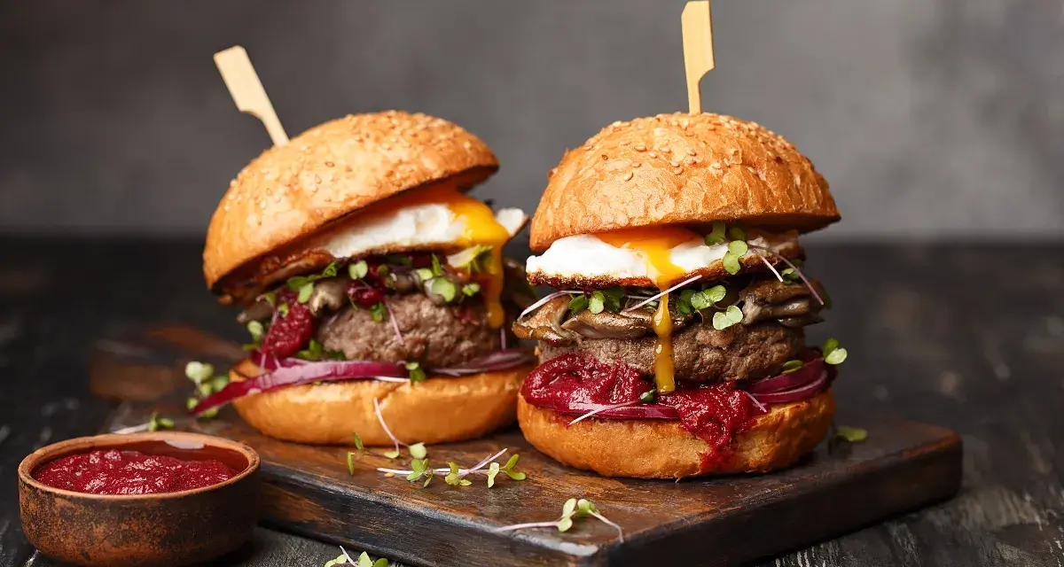Zwei saftige Burger mit Spiegelei, Pilzen und roten Zwiebeln, serviert auf einem Holzbrett. Ein Muss für jeden Burger Laden in Berlin.