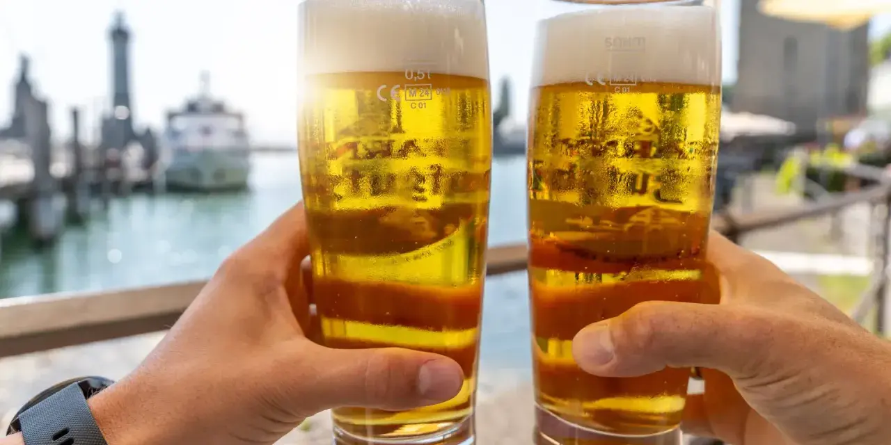 Zwei Hände halten Biergläser mit schaumigem Bier. Im Hintergrund ein Hafen. Ist Bier gesund? Ein Genuss am Wasser.