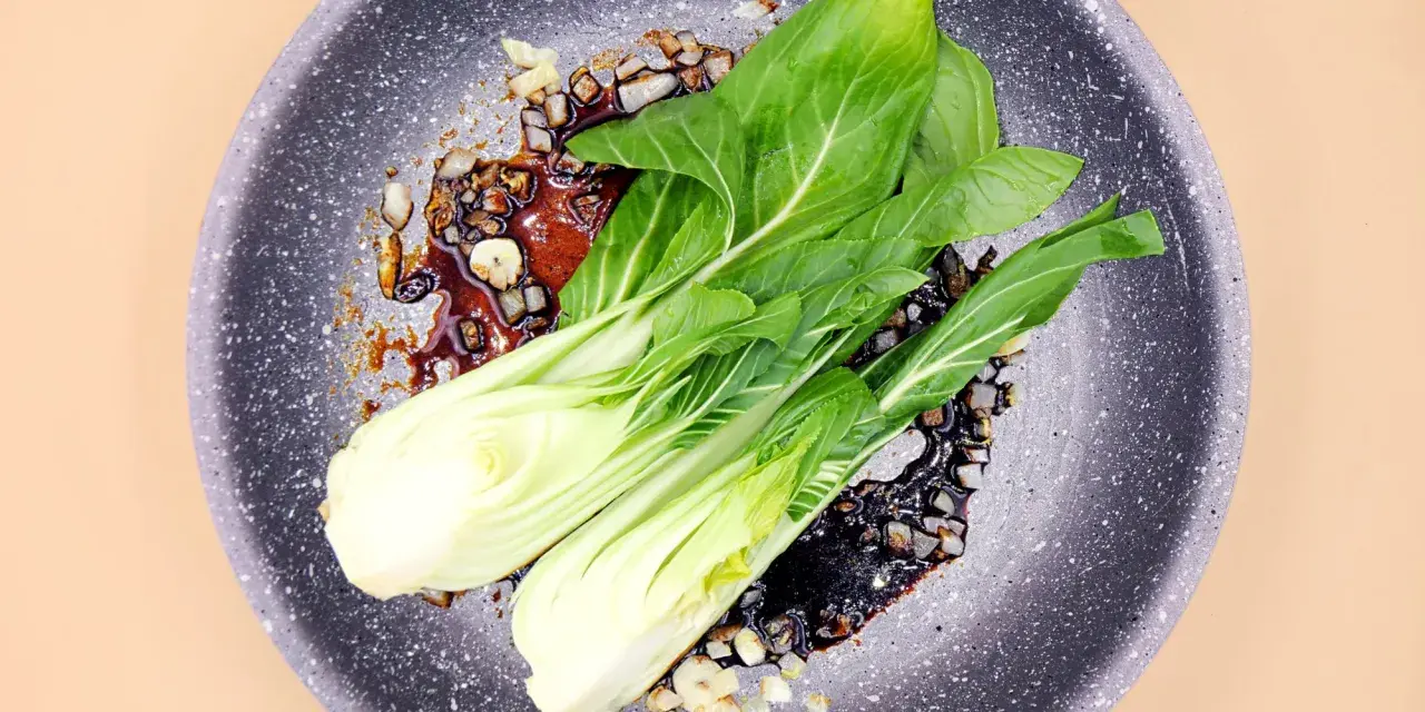 Na patelni smaży się aromatyczny sos z czosnkiem i cebulą, a na nim leży świeża sałata pak choi, gotowa do przygotowania pysznego dania.