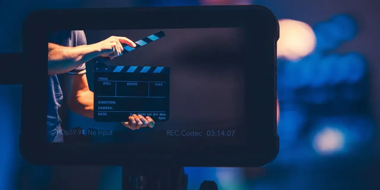Ręka zamyka klaps filmowy. Na ekranie widać napis "REC:Codec 03:14:07". To plan średni, zapowiadający rozpoczęcie nagrania.
