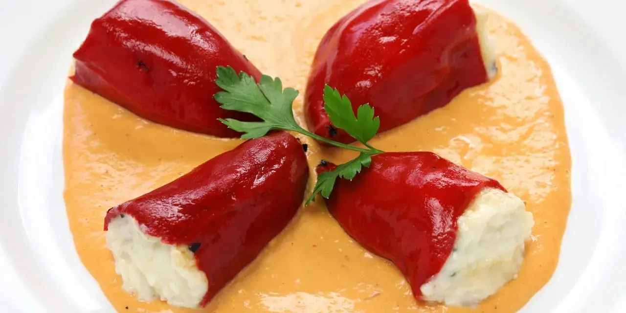 Cuatro pimientos rojos rellenos de bacalao, servidos sobre una cremosa salsa naranja y decorados con perejil.