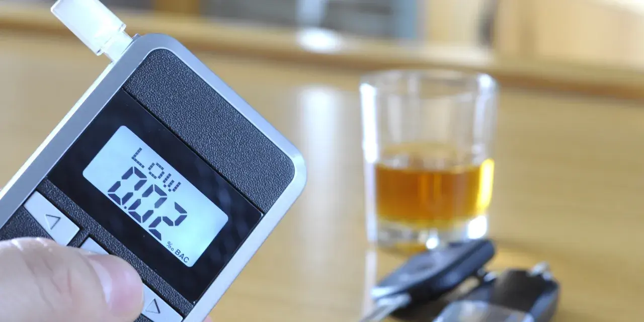 Alkomat z wyświetlaczem pokazującym 0.02% BAC, obok kieliszek z alkoholem i kluczyki samochodowe.