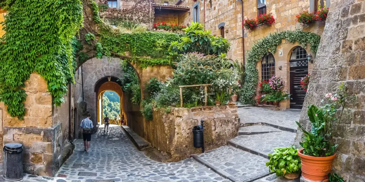 Un vicolo acciottolato con case in pietra ricoperte di edera e fiori. Un uomo cammina verso un arco che si apre su un paesaggio verde. Uno dei tanti posti belli nelle Marche.
