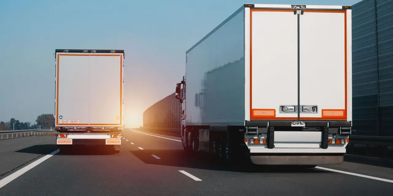 Dwa białe tiry jadą autostradą. Na jednym z nich widać znak zakazu wyprzedzania dla ciężarówek.