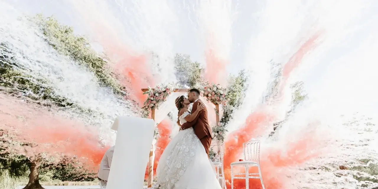 Un couple s'embrasse sous une arche fleurie, entouré de fumée rose et blanche. Ces photos mariage capturent un moment magique.