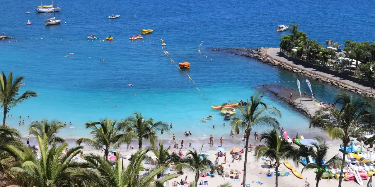 Słoneczne gran canaria plaże z palmami, turkusową wodą i ludźmi odpoczywającymi na piasku. Widać łodzie i sprzęt wodny.