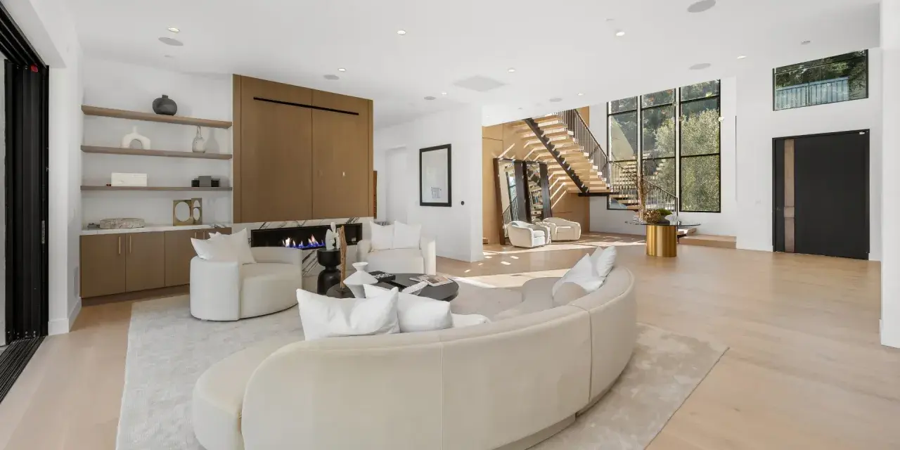 Soggiorno luminoso con camino moderno, divano curvo e poltrone bianche. Scale a vista e grandi finestre definiscono l'arredamento minimal chic.