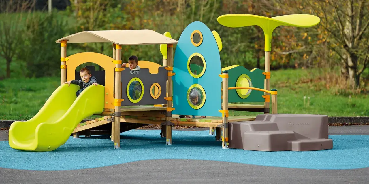 Des enfants jouent sur une aire de jeux colorée, respectant la réglementation aire de jeux extérieur. Un toboggan vert vif et une structure inspirée d'une fusée bleue.