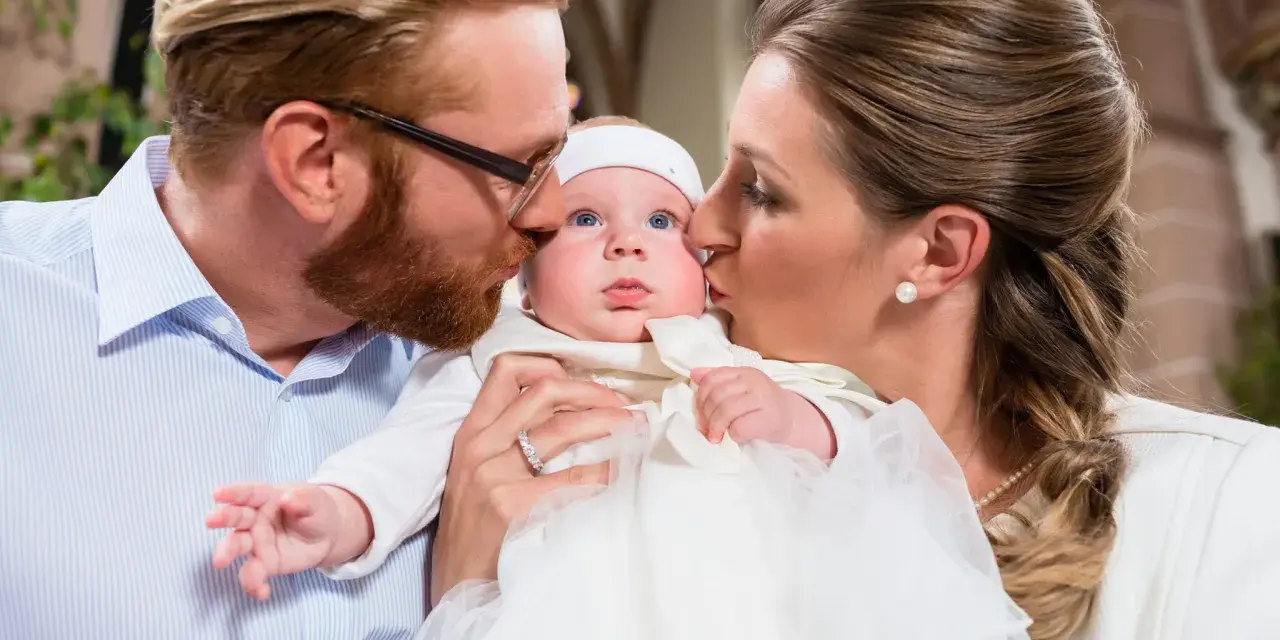 Eltern küssen ihr Baby bei der Taufe. Die Frage, wie viele Paten darf man haben, beschäftigt viele.