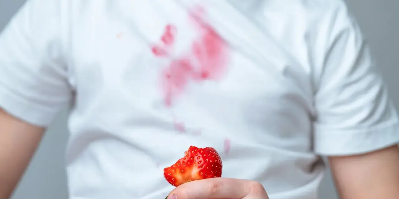 Dziecko trzyma kawałek truskawki, a na białej koszulce widać czerwone plamy z truskawek.