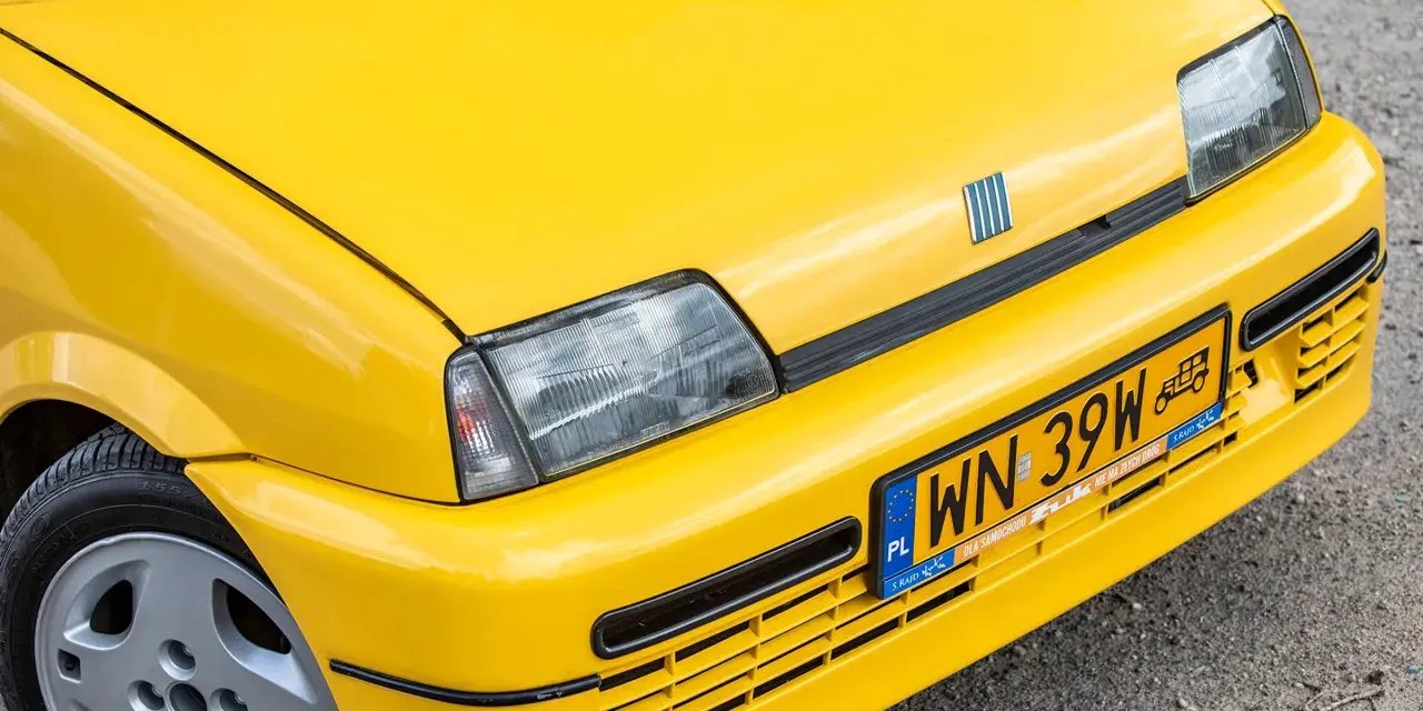 Żółte tablice rejestracyjne na błyszczącym, żółtym samochodzie Fiat. Widoczny fragment maski, reflektor i zderzak.